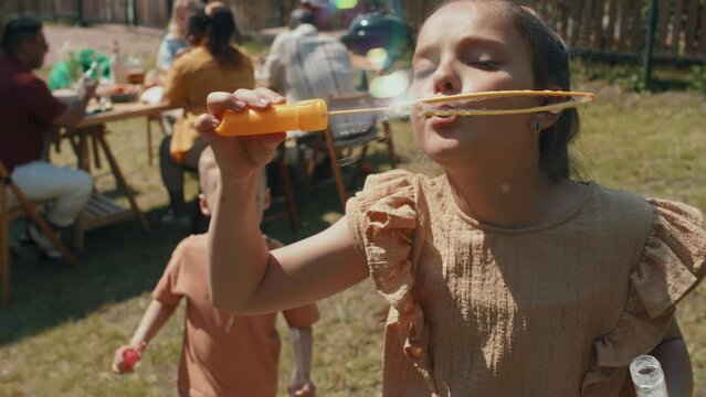 Slow motion of joyful preteen girl blowing soap bubbles outdoors on summer day during backyard bbq party