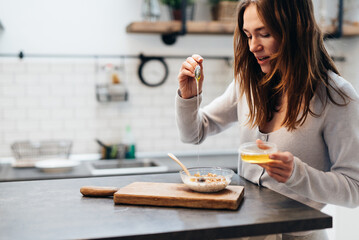 Young woman in the kitchen adds honey to granola