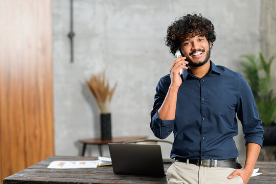 Happy Inspired Indian Male Small Business Owner Talking On The Smartphone While Standing In Modern Loft Office. Smiling Successful Contemporary Man Manager In Casual Wear Holding Phone Call,copy Space