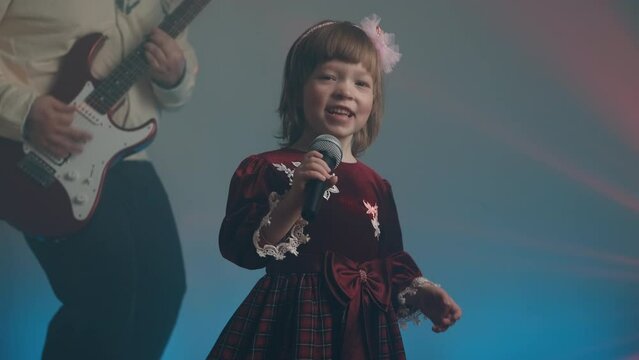 Little girl on stage in vintage dress, she sings into microphone and dances, her father plays an electric guitar. Color music is shining and smoke is billowing. Family time, creativity and hobbies