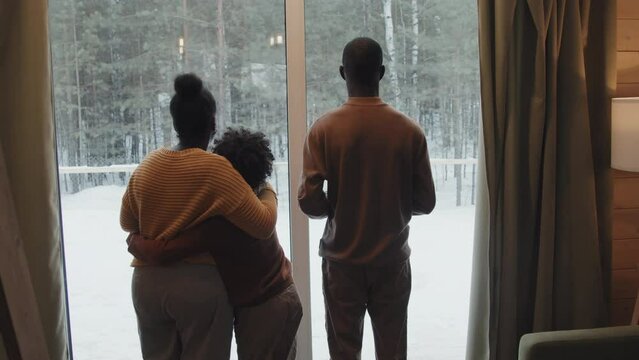 Rear view of Black boy standing in front of window together with his parents enjoying beautiful view of pine woods on winter day