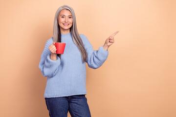 Photo of funny pretty retired woman wear blue sweater drinking coffee pointing empty space isolated beige color background