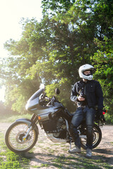 Fototapeta premium rider guy use smart phone in jeans biker jacket and helmet sit on tourist touring motorcycle. outdoors, dual sport adventure concept, vertical photo, dirt road