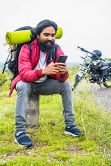 vertical shot of Happy smiling traveler busy using mobile phone while sitting on road side infrtont of motor bike - concept of technology, social media and taking break.