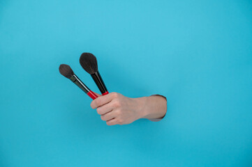 A woman is holding two large brushes for cosmetics. Woman's hand sticking out of blue paper background. 