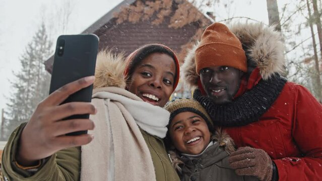 Stylish African American family of three wearing warm clothes standing outdoors against their country house on forest village taking selfies on snowy day