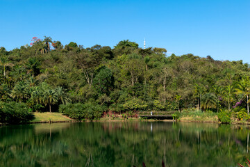 Linda vista com algumas construções artísticas, muita vegetação ao redor, lindo céu azul com nuvens e um grande lago em frete, refletindo a construção e o céu, em museu a céu aberto de Minas Gerais.