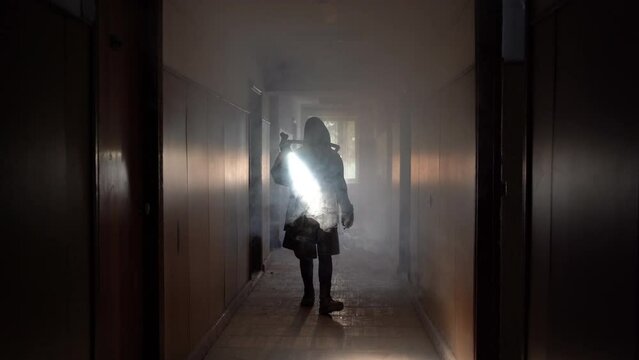Woman in gas mask and cloak in smoke with flashlight an building at night, air pollution