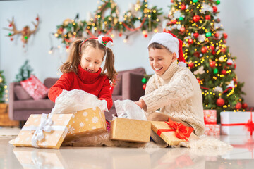 cute cheerful children with an open smile, brother and sister, open golden gift boxes tied with red ribbon. children laugh and hold gifts in their hands. Christmas tree in light garlands in background