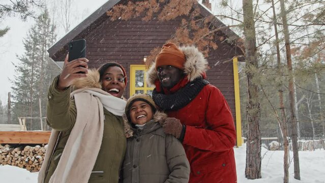 Slow motion of modern African American family with kid standing outdoors against brown wooden country house taking selfie photos on winter day