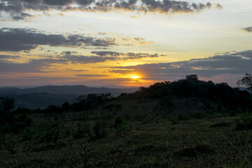 Lindo pôr do sol entre nuvens e silhueta de árvores ao ao longe, entardecer com céu dourado, de cima de monte no bairro Jardim das Oliveiras, Esmeraldas, Minas Gerais, Brasil.