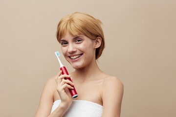 a joyful blonde woman with her hair pinned back is standing on a beige background wrapped in a towel and holding a red electric toothbrush in her hand, smiling and squinting her eyes with delight