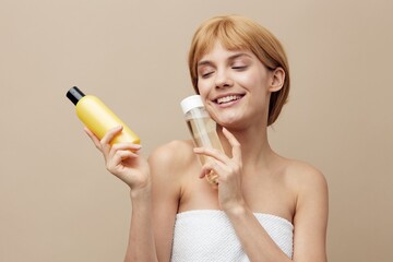 Fototapeta premium a happy, friendly woman with clean skin, stands on a beige background and advertises jars of cosmetics in her hands, smiling with her eyes closed, lifting them to her face.