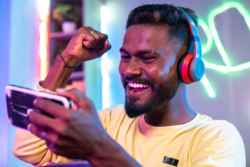 Excited gamer with headphones celebrating win game by raising hands while playing live video game on mobile phone at home - concept of successful, gaming addiction and tournament © WESTOCK