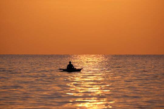 Dark silhouette of lonely fisherman rowing on his boat on sea water at sunset