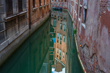Reflections of the sky and houses on the surface of the water of a narrow canal street in Venice, old walls of houses with windows on a Venetian street, a lonely boat near the brick wall of the house