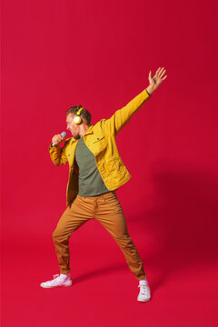 Singing young man enjoying song dancing with one hand up using phone and wireless headphones wearing jeans yellow jacket isolated on red background. Man sing while recording his voice