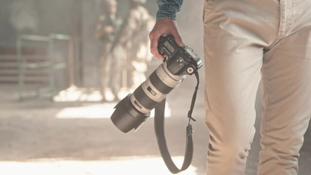 Close up of man hand holding professional photo camera. War journalist walking in old ruined building carrying camera in hands. Armed soldiers on background. War press. Dangerous work. Sensation