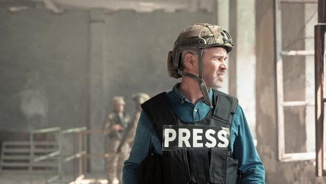 Close up portrait of middle-aged tired war journalist wearing helmet and bulletproof vest walking in old ruined building looking at window. Armed soldiers on background. War press correspondent