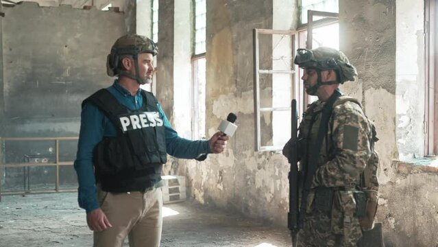 Male war journalist in bulletproof vest and helmet reporting live in destroyed building conducting interview with military representative Caucasian man correspondent making reportage for media channel