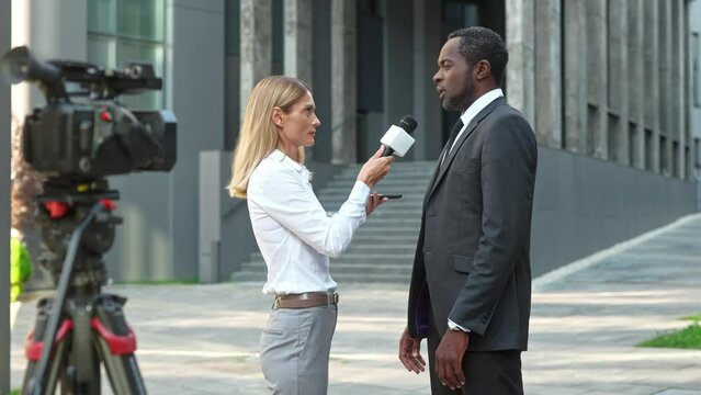 Portrait of beautiful Caucasian female TV reporter holding smartphone and microphone interviewing African American male politician outdoor in street. Journalist live reportage. News. Politics concept