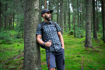 Naklejka na ściany i meble Hiker beard man in a green mountain forest. Dressed up in a cap, shorts, and plaid shirt with a backpack and trekking sticks
