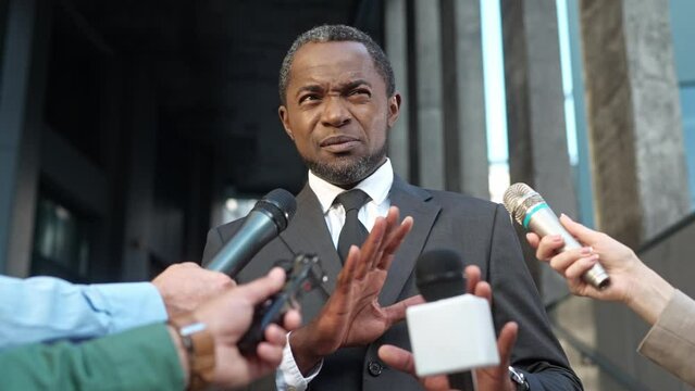 Close up of male public relations representative answering questions giving interview standing outdoors. Handsome serious politician crowded by many news journalists with microphones in street
