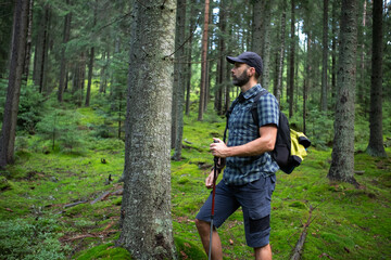 Hiker beard man in a green mountain forest. Dressed up in a cap, shorts, and plaid shirt with a backpack and trekking sticks