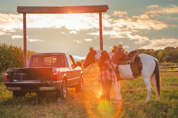 A cowgirl saddles a horse at a ranch. Man, horse and pickup truck in the field at sunset. - Powered by Adobe