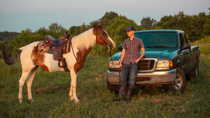 A cowboy on a ranch near a pickup truck. Western horse on the field at sunset. Cowboy saddle and boots. - obrazy, fototapety, plakaty