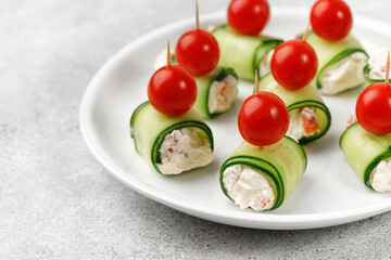 Cucumber canapes, rolls stuffed with cream cheese, sweet pepper, tomatoes cherry over it, served on a white plate. Holiday vegetable appetizer, snack for a festive table.