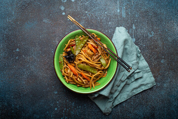 Asian dish stir fry udon noodles with chicken, vegetables and mushrooms in green ceramic bowl with wooden chopsticks on rustic dark blue concrete background from above, Chinese or Thai fast food 