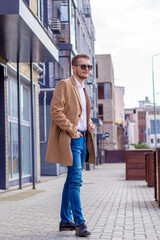 A young man dressed in a beige coat is walking down the street. A young bearded guy with a modern hairstyle and sunglasses on an urban background