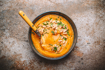 Ceramic bowl with pumpkin cream soup decorated with peanuts on rustic old metal oven tray background with spoon, from above. Autumn cozy dinner concept 
