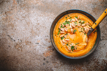 Ceramic bowl with pumpkin cream soup decorated with peanuts on rustic old metal oven tray background with spoon, from above. Autumn cozy dinner concept 

