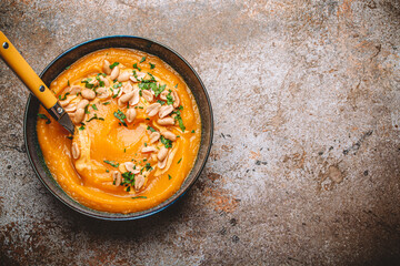 Ceramic bowl with pumpkin cream soup decorated with peanuts on rustic old metal oven tray background with spoon, from above. Autumn cozy dinner concept 
