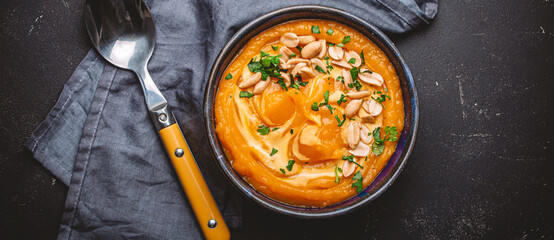 Ceramic bowl with pumpkin cream soup decorated with peanuts on black rustic stone table background with spoon, from above. Autumn cozy dinner concept, space for text

