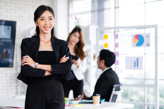 Portrait of a smiling professional Asian businesswoman wearing a black blazer standing in front of blur colleagues meeting in the background. Entrepreneur leadership image with copy space.