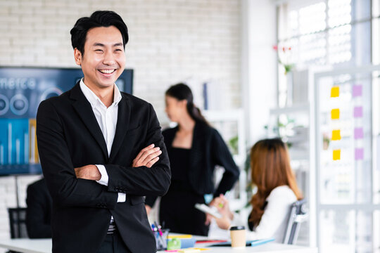 Portrait of a smiling professional Asian businessman wearing a black suit standing in front of blur colleagues meeting in the background. Entrepreneur leadership image with copy space.