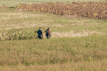 Two hunters armed with rifles wait to shoot their prey in a field with dried sunflowers in the background