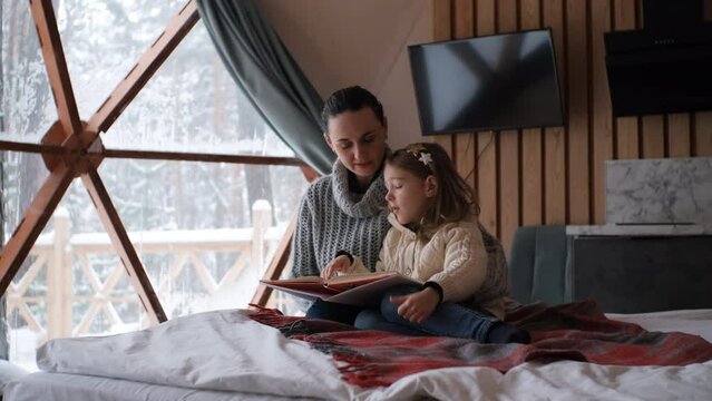 Young woman with her cute little daughter reading book while sitting in bed of dome camping. Winter glamping vacation and travel concept