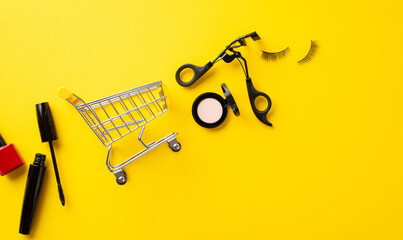 Black friday sales concept. Top view photo of shopping cart cosmetics mascara false eyelashes eyeshadow nail polish and lashes curler on isolated yellow background