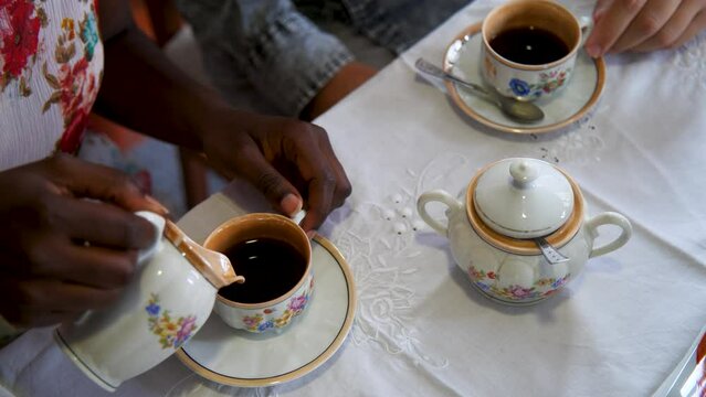 Close up of interracial young couple's hands preparing and drinking a coffee with milk and sugar using a vintage porcelain cup with flowers pattern.