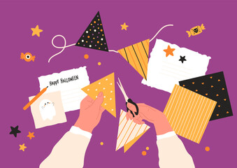 Halloween preparation concept, hands cut out flags for bunting with scissors. Flat lay with craft paper, greeting card with cute ghost, happу Halloween text, pencil, candy,  stars on purple background