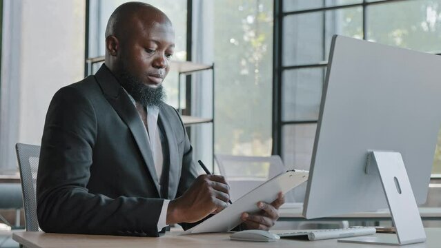 African writer writing notes on paper tablet sit at office desk adult entrepreneur american businessman create new ideas corporate tasks business strategy marketing plan fill out points in checklist