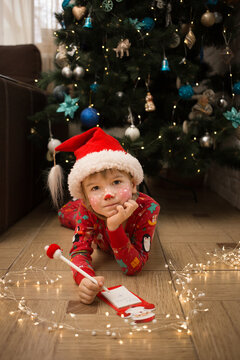child in Christmas pajamas, Santa's hat and deer make-up writes letter to Santa Claus lying on floor in front of Christmas tree. winter holidays, interesting childhood. Cozy festive atmosphere at home