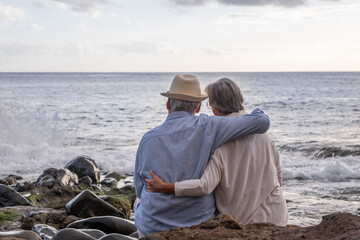 Rear view of relaxed caucasian senior couple sitting on the pebble beach at sunset light admiring...