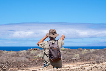 Rear view of active senior man with hat and backpack in outdoor excursion looking at the horizon over sea and pointing at the big cloud in front to him