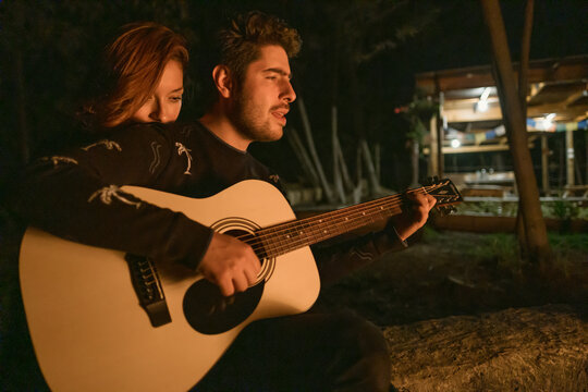 beautiful couple playing guitar around a campfire in the evening