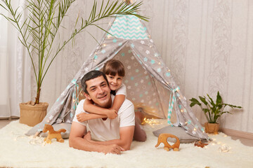 Image of smiling handsome Caucasian man wearing white t shirt lying on floor with his little daughter near her wigwam, cute child hugging her father and expressing happiness. © sementsova321
